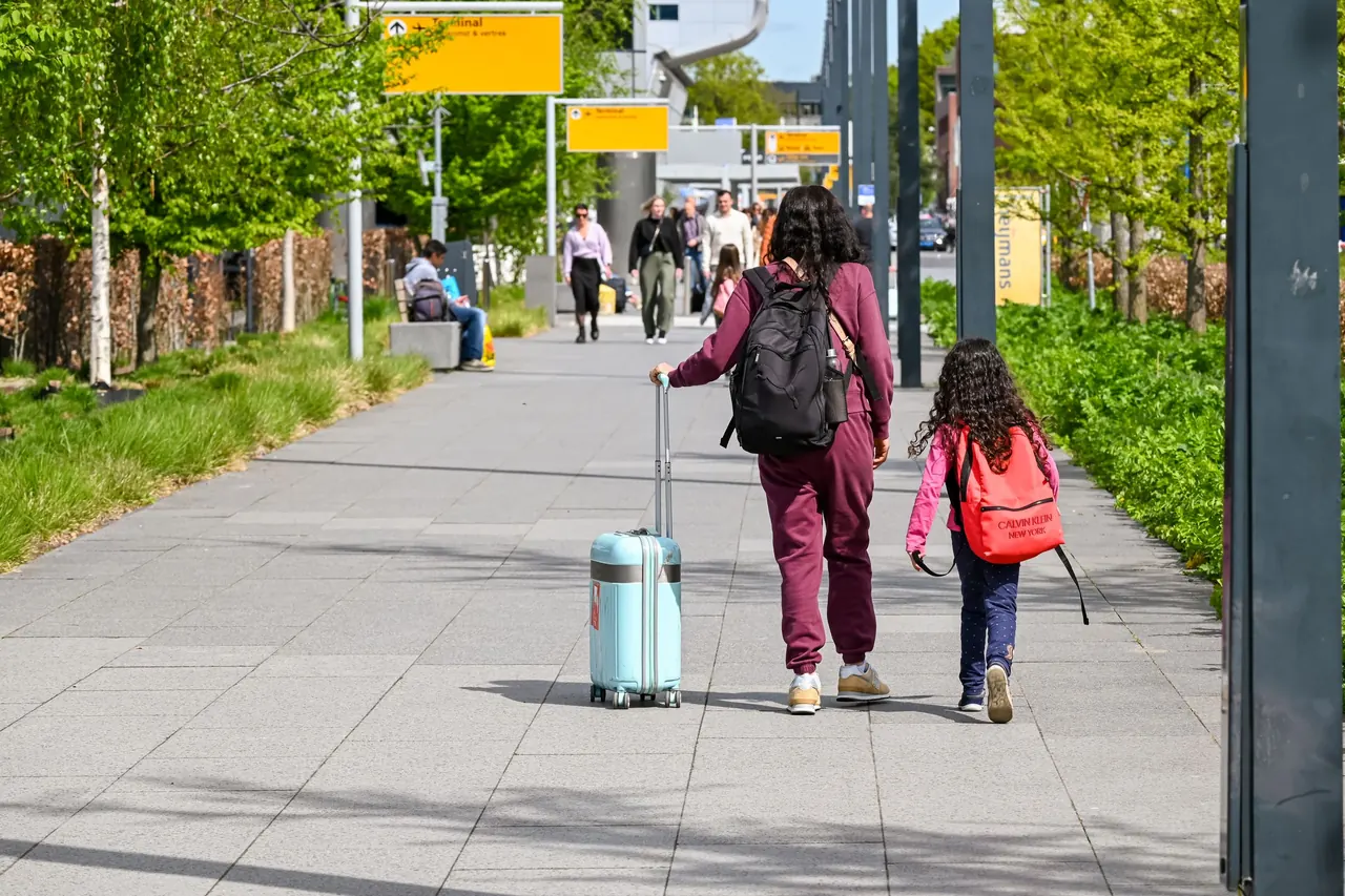 A mother and daughter walking with luggage outside Eindhoven Airport on a sunny day.